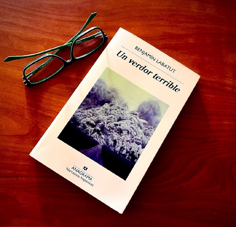Portada del libro 'Un verdor terrible' de Benjamín Labatut (Anagrama) sobre una mesa de madera, junto a unos espejuelos