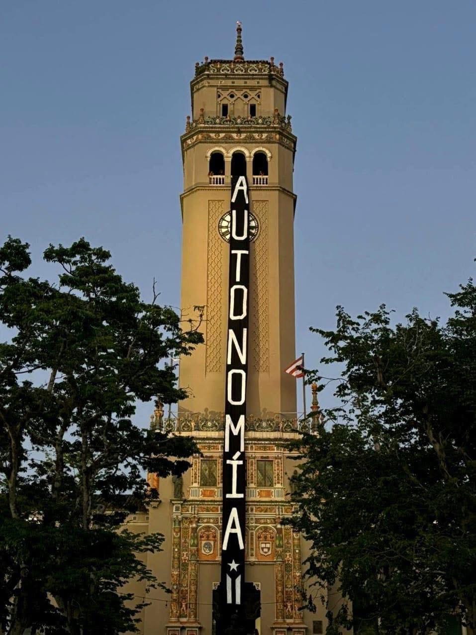 La torre del reloj de la Universidad de Puerto Rico en Río Piedras con una pancarta vertical negra que lee 'AUTONOMÍA'