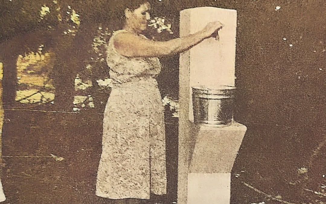 Fotografía antigua en sepia de una persona de pie junto a una estructura de concreto, posiblemente relacionada con el suministro de agua