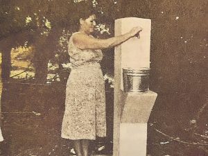Fotografía antigua en sepia de una persona de pie junto a una estructura de concreto, posiblemente relacionada con el suministro de agua